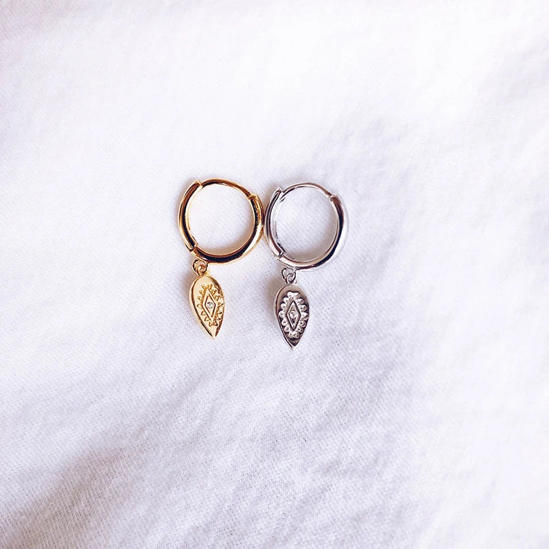 A pair of small hoop earrings with teardrop leaf charms, one in gold and one in silver, displayed on a textured white fabric background.
