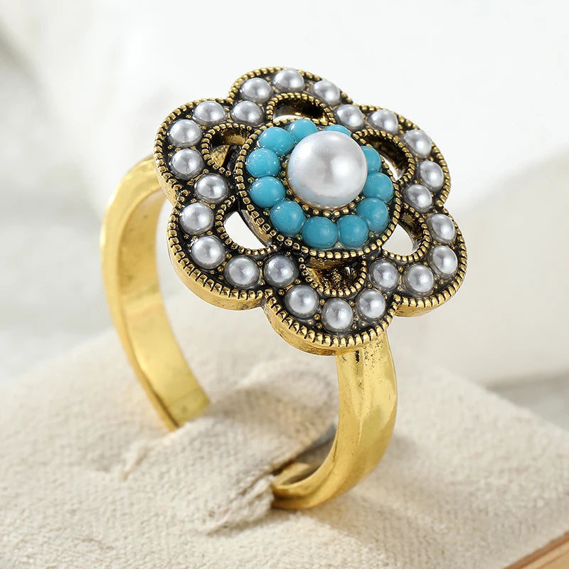 A gold ring with a floral design, featuring a central white pearl surrounded by turquoise blue beads, and outer silver-gray faux pearls, displayed on a beige ring stand.