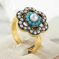 A gold ring with a floral design, featuring a central white pearl surrounded by turquoise blue beads, and outer silver-gray faux pearls, displayed on a beige ring stand.