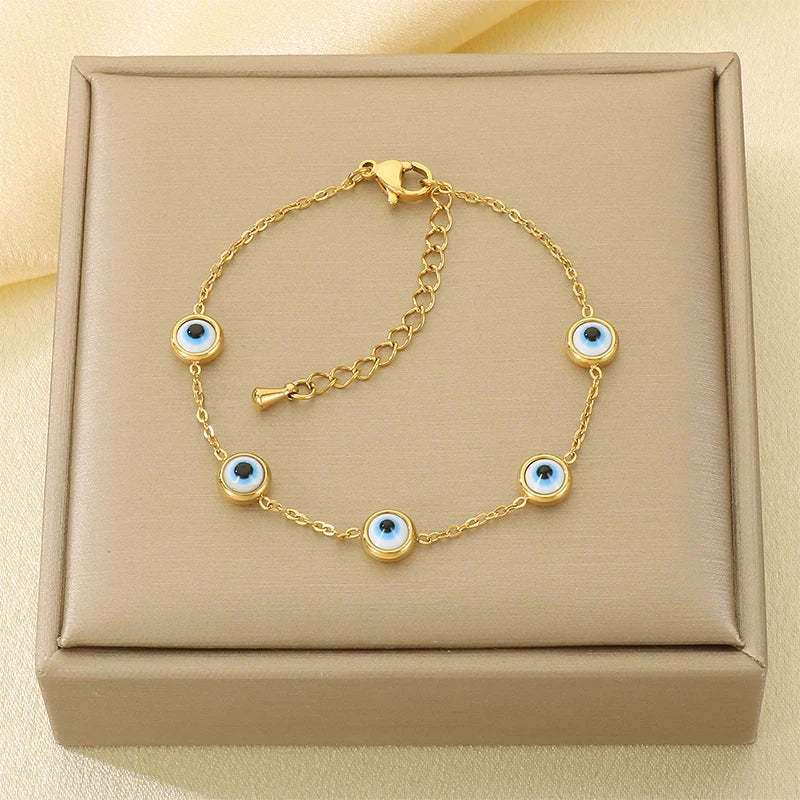 A gold chain bracelet with five evil eye charms, displayed on a beige jewelry box.