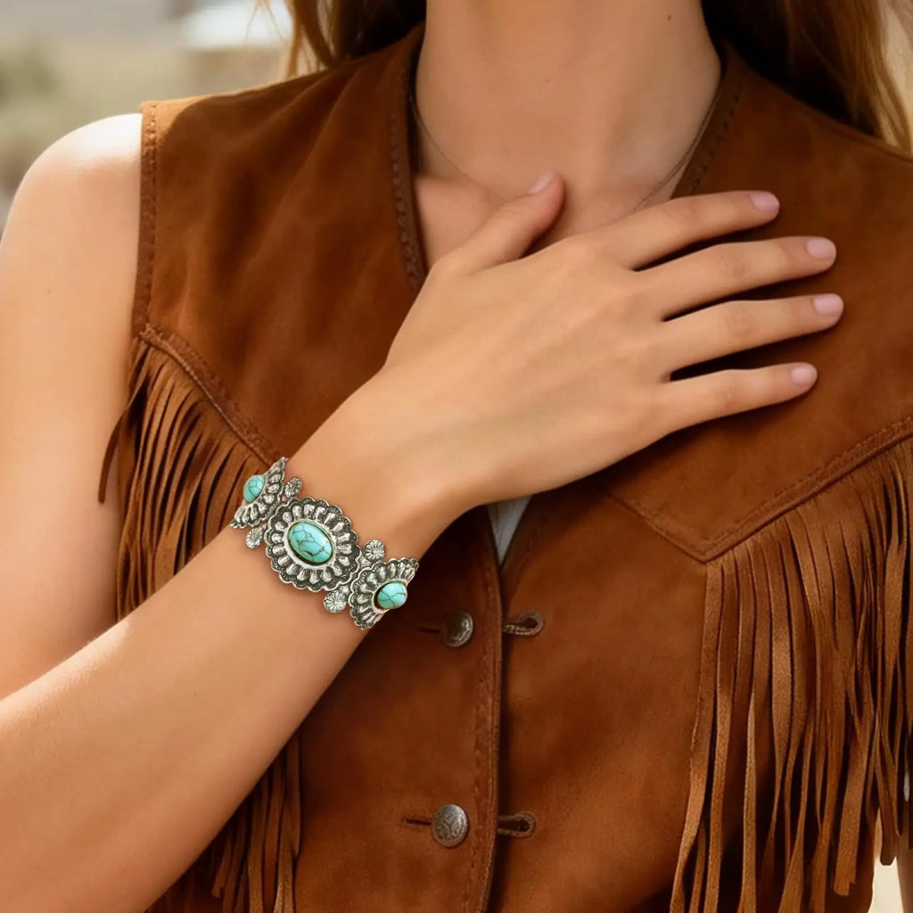 A close-up of a woman wearing a wide silver-toned bracelet with turquoise stones, paired with a brown suede fringe vest.