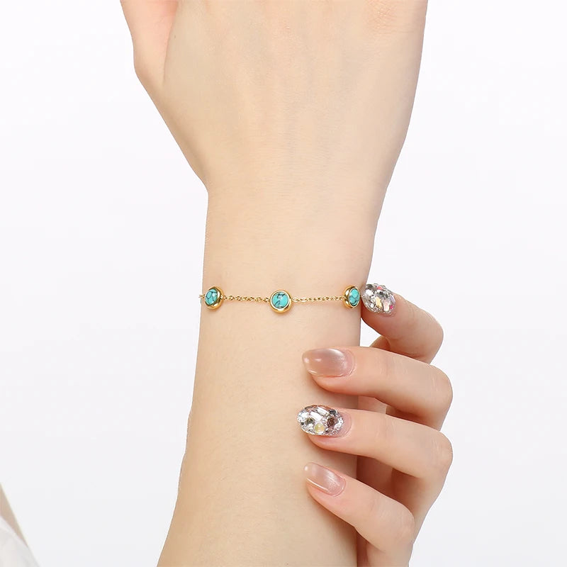 A delicate gold chain bracelet with small turquoise blue oval stones, shown worn on a woman's wrist with manicured nails featuring rhinestone nail art.