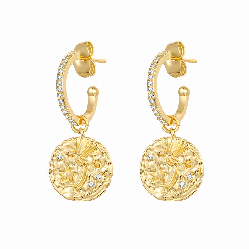 A pair of gold huggie hoop earrings with small clear pavé stones along the front and a dangling round coin-style medallion featuring an embossed profile and small clear stones, shown on a plain white background.