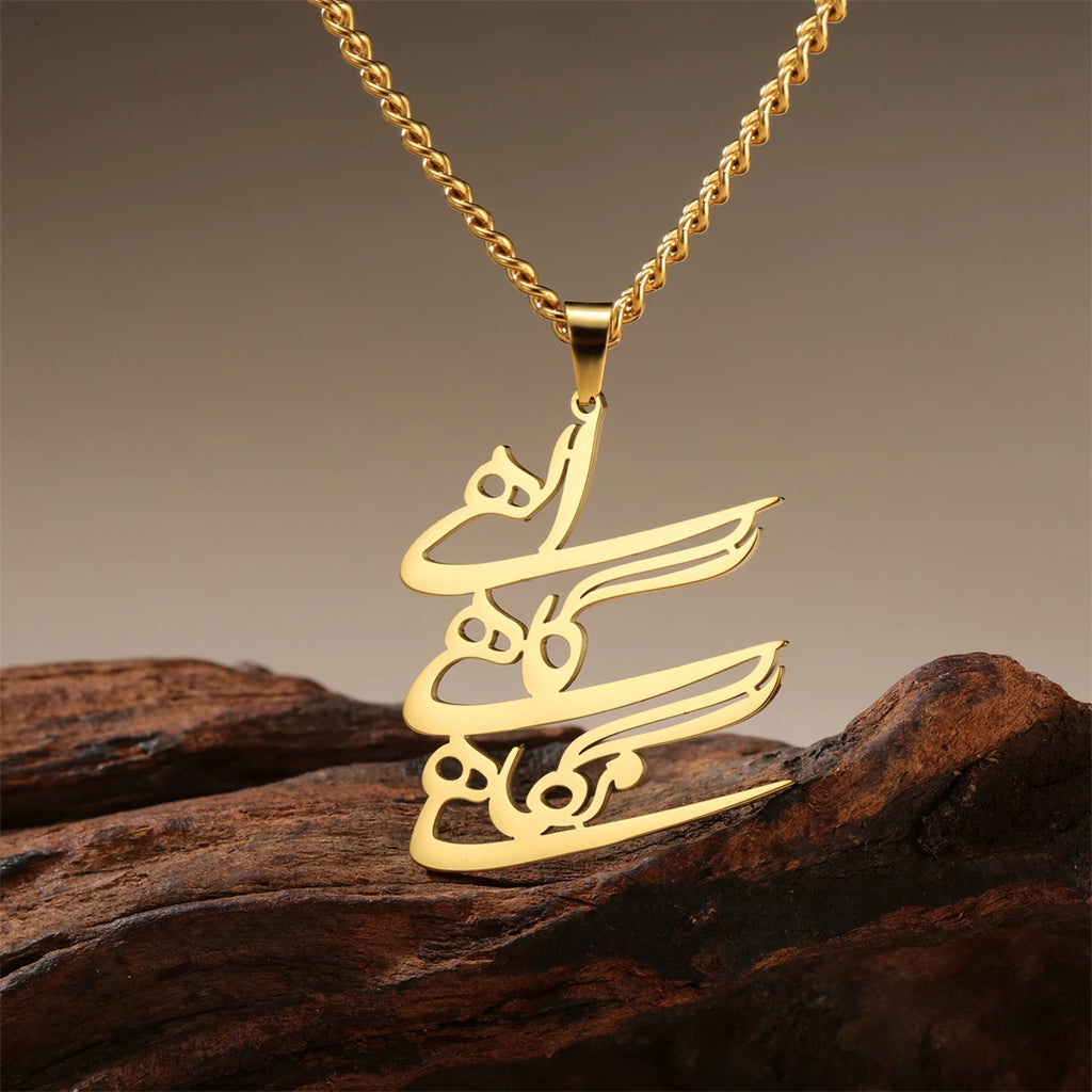 A gold-tone necklace with a calligraphy pendant resting on a piece of dark wood against a warm gradient background.
