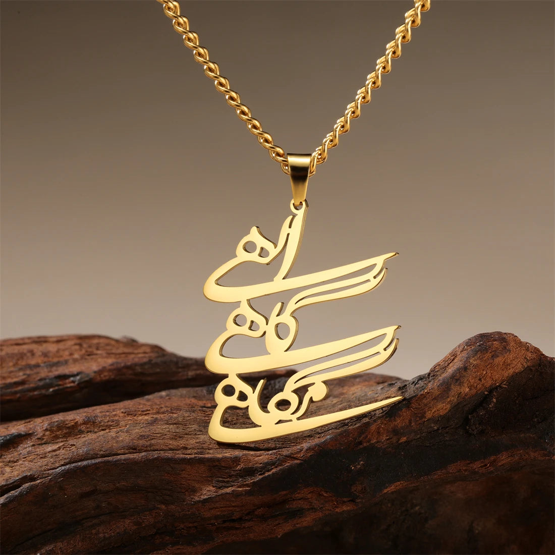 A gold-tone necklace with a calligraphy pendant resting on a piece of dark wood against a warm gradient background.