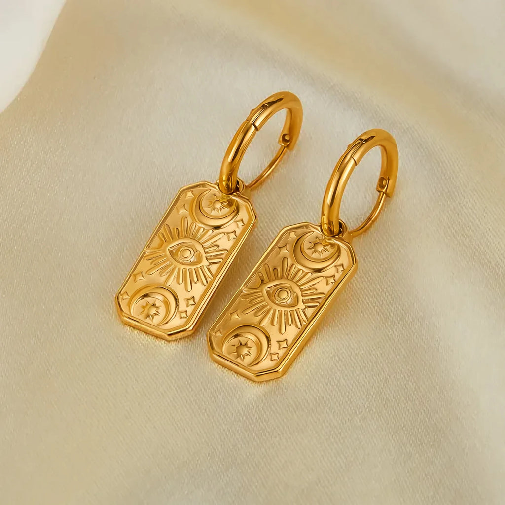 Gold evil eye earrings with intricate designs on beige fabric background