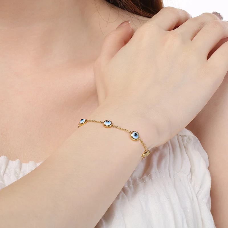 A delicate gold chain bracelet with three evil eye charms, worn on a woman's wrist. The bracelet features light blue and white eye designs set in gold.
