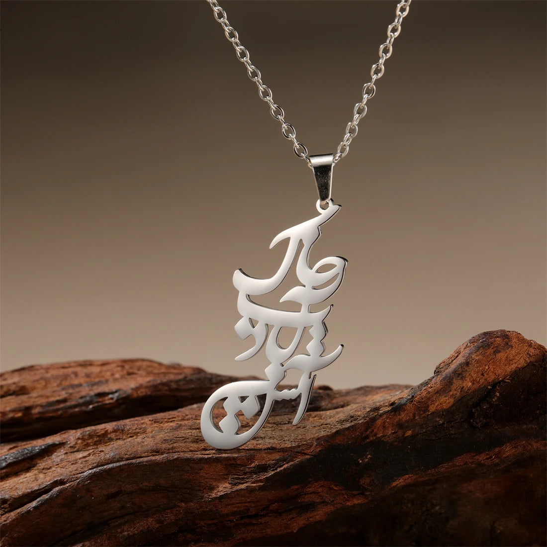 A silver-toned metal pendant necklace with an intricate calligraphic design, resting on a textured piece of dark brown wood against a warm brown gradient background.