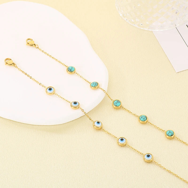 A pair of gold chain bracelets with small evil eye charms, displayed on a white curved display stand against a light beige background.