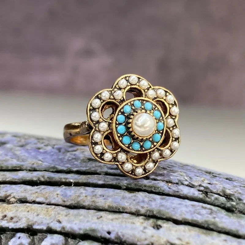An ornate vintage-style gold ring with a flower-shaped setting, featuring a central white pearl surrounded by turquoise and small white stones, resting on a textured gray stone surface.