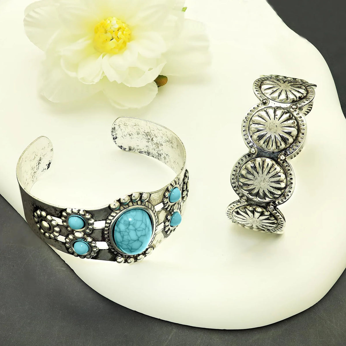 Two silver-toned cuff bracelets with turquoise stones, displayed on a white sculptural surface with a white flower prop. One bracelet is a wide open cuff with a large oval turquoise center and smaller round turquoise accents; the other is a slim matching cuff with stacked oval turquoise motifs.