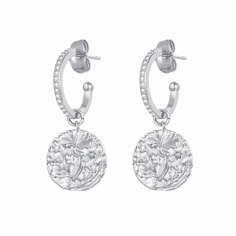 A pair of silver huggie hoop earrings with small clear stones along the front and a dangling round coin-style medallion featuring an embossed profile.