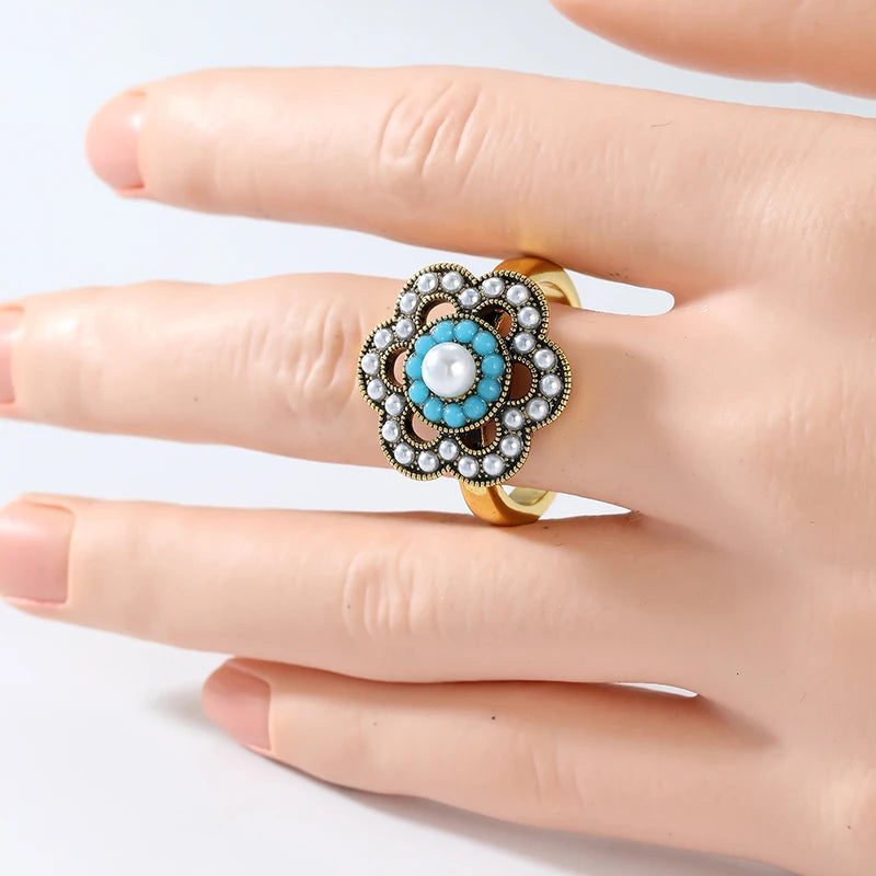 A gold ring with a turquoise and white pearl floral design, shown worn on a hand.