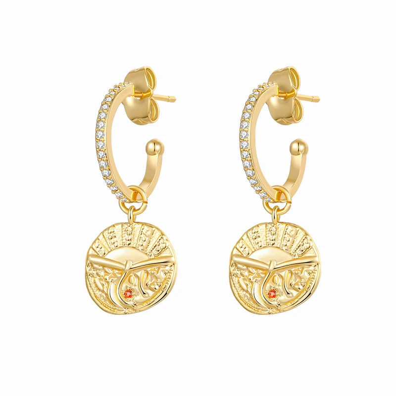 A pair of gold hoop earrings with a circular sunburst pendant featuring a central dagger motif and a small red gemstone, accented by a row of clear stones along the upper curve, set against a plain white background.