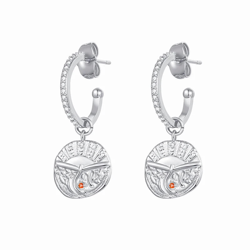 A pair of silver hoop earrings with a circular medallion charm featuring a cross and a small orange stone, shown on a plain white background.