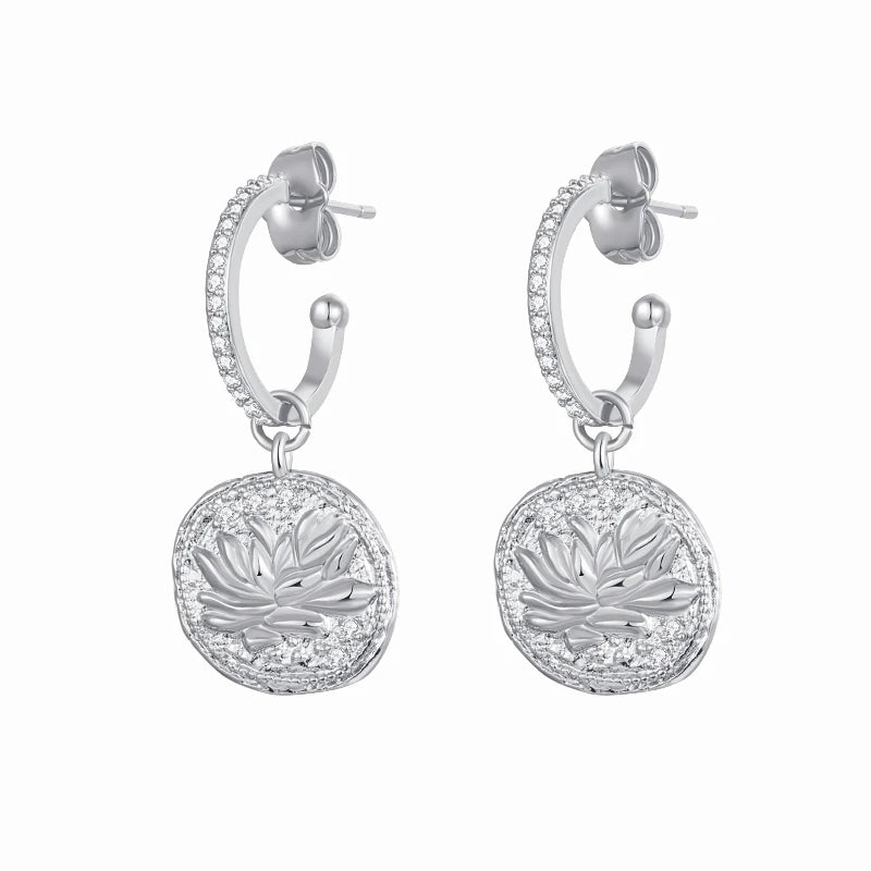 A pair of silver huggie hoop earrings with pavé-set clear stones and a dangling round medallion featuring an engraved lotus flower, shown on a plain white background.