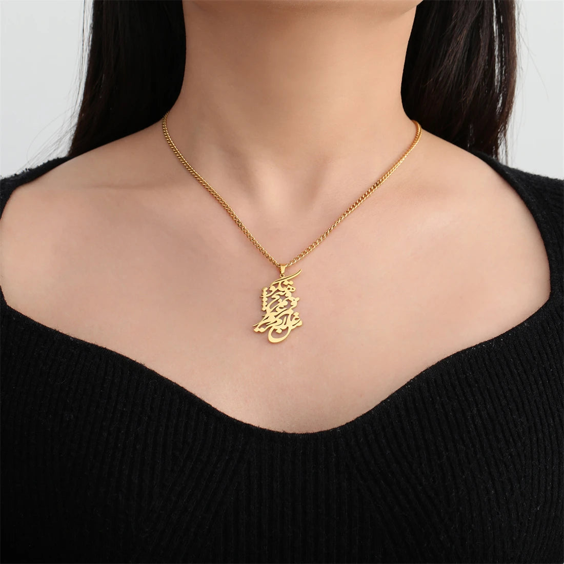 A gold chain necklace with a detailed Arabic calligraphy pendant is shown on a model's neck, wearing a black ribbed top against a light gray background.