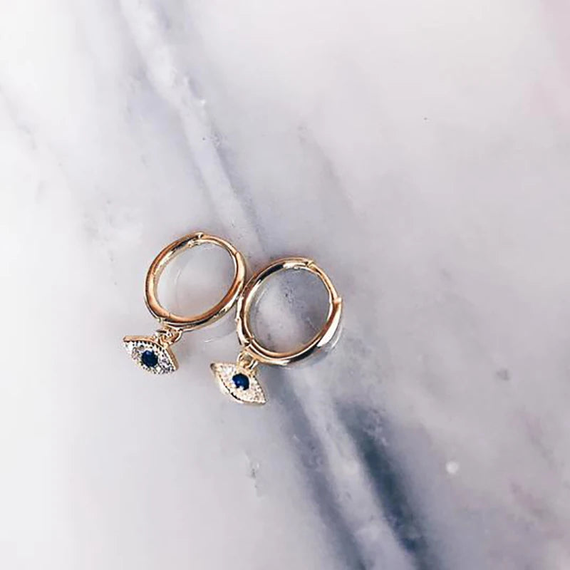 A pair of gold huggie hoop earrings with dangling evil eye charms featuring blue and clear stones, displayed on a light gray marble surface.