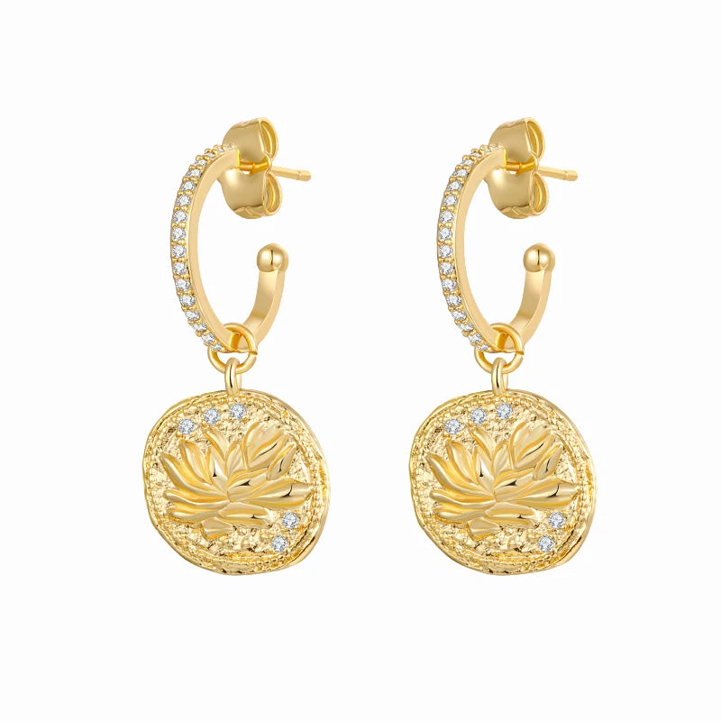 A pair of gold huggie hoop earrings with pavé-set clear stones and round textured gold disc charms featuring a lotus flower design, shown on a plain white background.