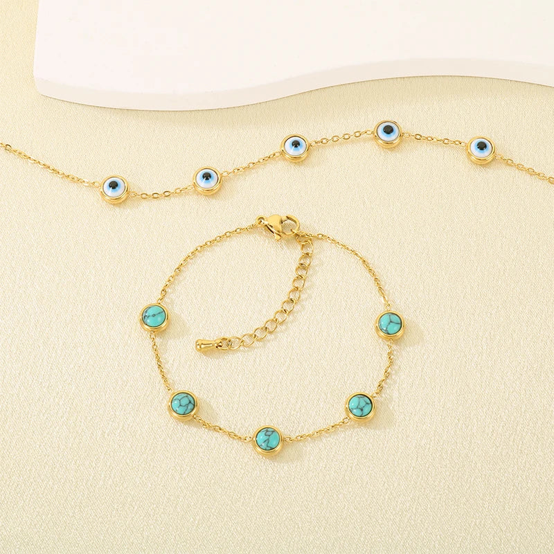 Gold-tone chain necklace and matching bracelet, each decorated with multiple round evil eye charms featuring turquoise-blue centers and white details, displayed on a light cream textured surface.