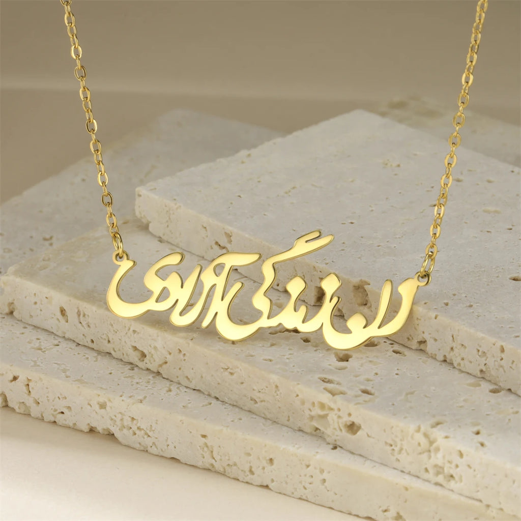 A gold-tone necklace with an Arabic nameplate pendant, displayed on layered beige stone tiles.