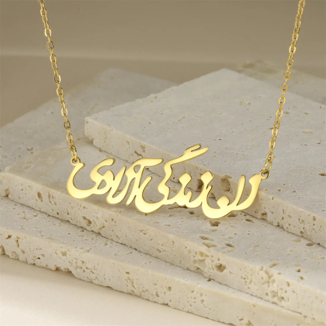 A gold-tone necklace with an Arabic nameplate pendant, displayed on layered beige stone tiles.