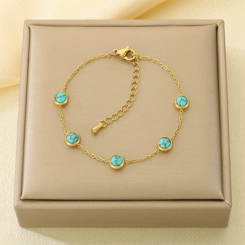 A gold chain bracelet with five round turquoise-blue stone accents, displayed on a beige jewelry box against a soft cream fabric background.