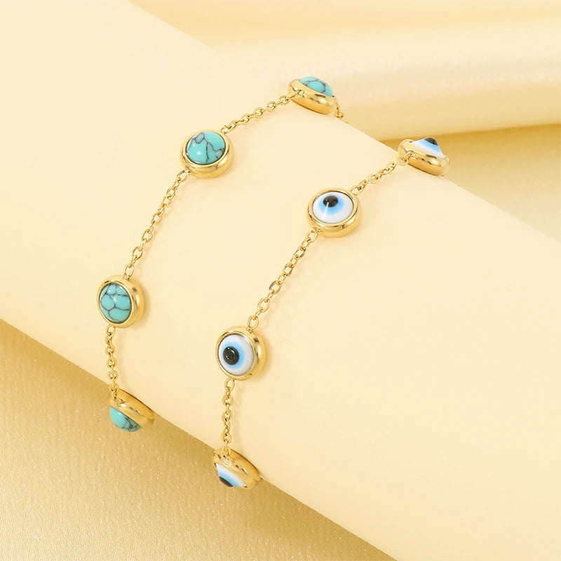 Gold chain bracelets with turquoise and evil eye charms, displayed on a cream-colored wrist form.