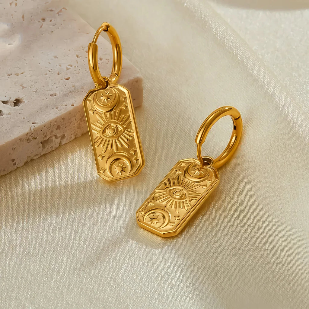 Gold evil eye earrings with intricate designs on beige fabric background