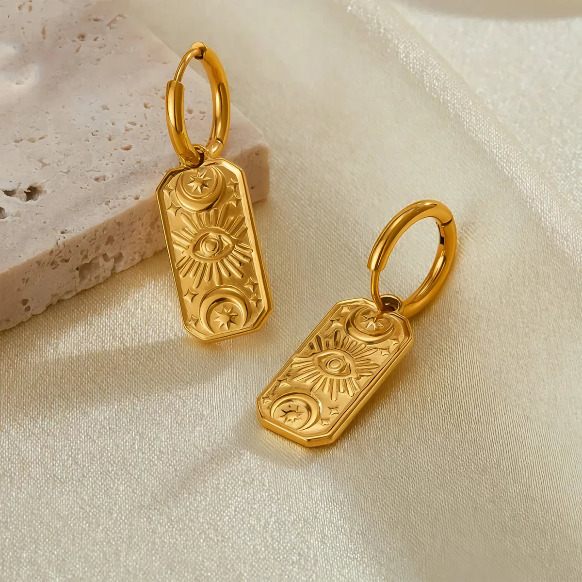 Gold evil eye earrings with intricate designs on beige fabric background