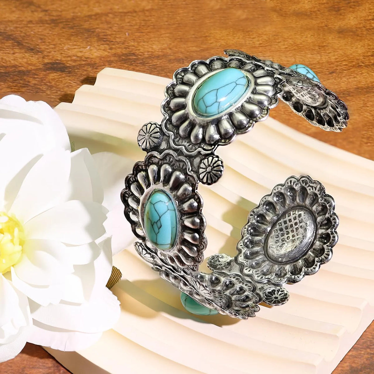 A wide silver-toned cuff bracelet with multiple oval turquoise stones and ornate floral engraved details, displayed on a cream-colored book or stand beside a white flower on a wooden surface.