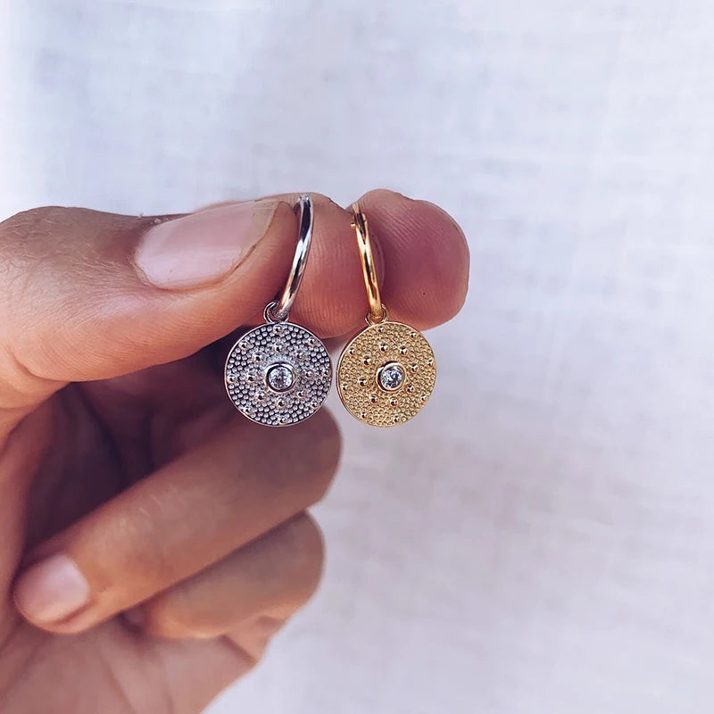 Two gold earrings with circular designs held between fingers against a white background