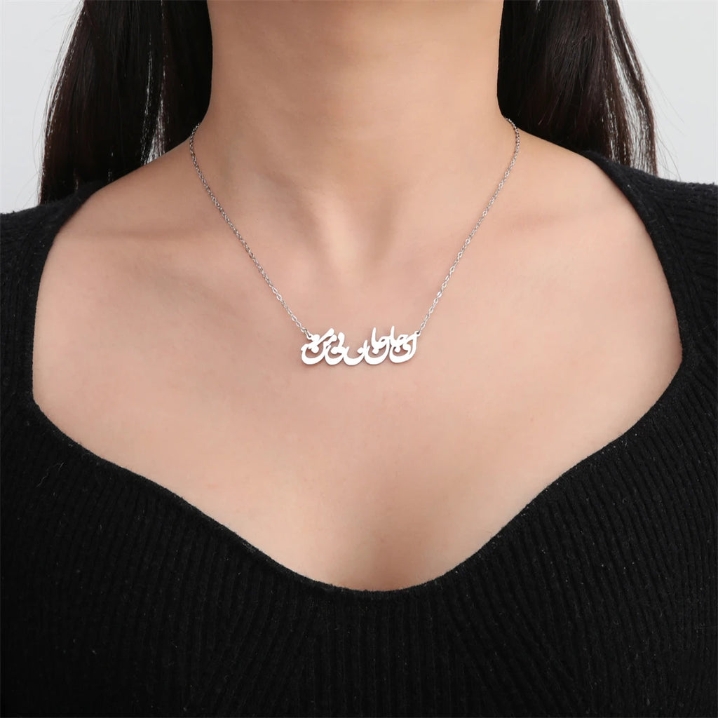 A close-up of a model wearing a delicate silver chain necklace with a custom Arabic calligraphy pendant spelling 'حبا المنزل'. The model is wearing a black ribbed knit top against a clean, light gray background.