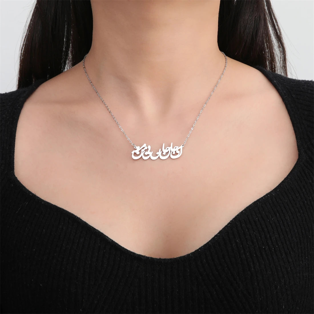 A close-up of a model wearing a delicate silver chain necklace with a custom Arabic calligraphy pendant spelling 'حبا المنزل'. The model is wearing a black ribbed knit top against a clean, light gray background.