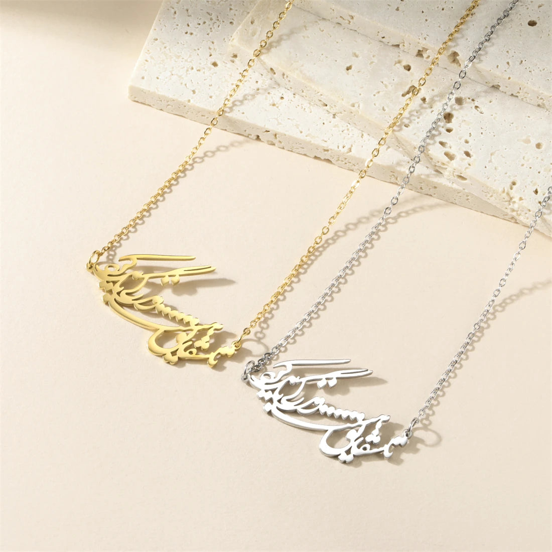 Two personalized nameplates on chains, one in gold and one in silver, displayed on a light beige surface with a textured stone prop.