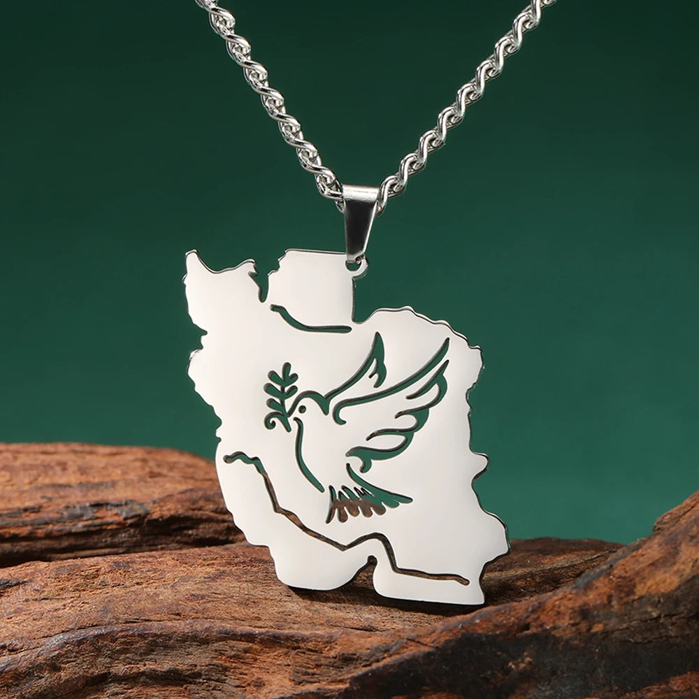 A silver-toned pendant necklace shaped like the map of Iran, featuring a green dove and olive branch design, displayed on a piece of rustic wood against a dark green background.