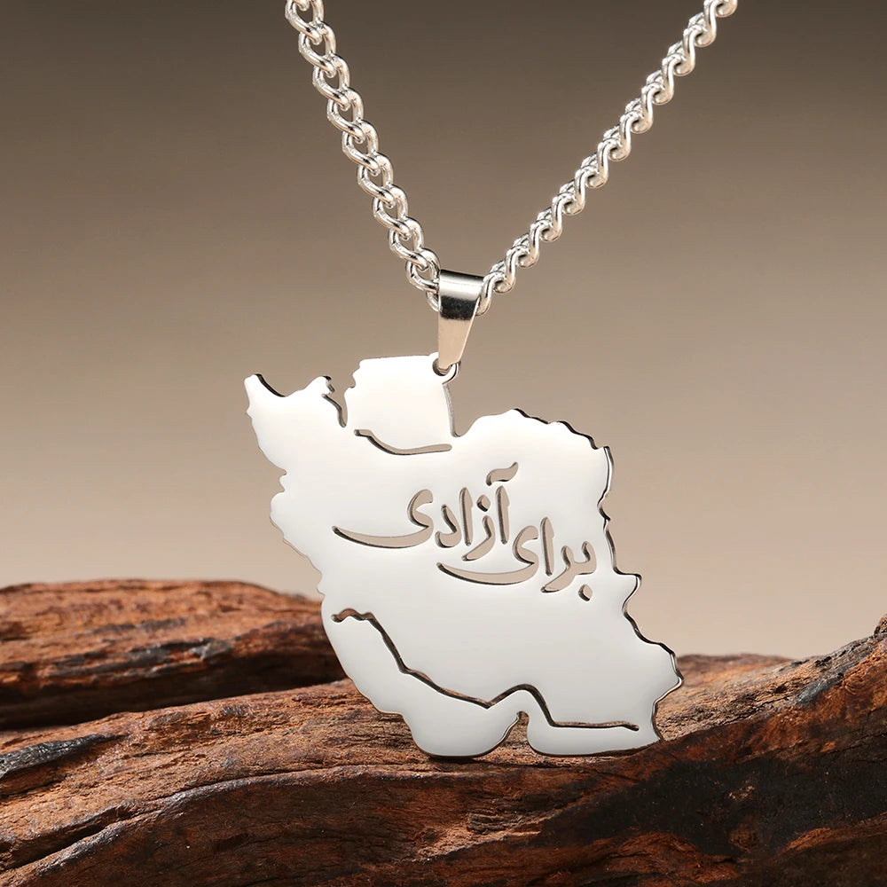 A silver-tone necklace with a pendant shaped like the map of Iran, featuring the Persian text 'برو آذایم' cut out in the center, resting on a piece of weathered wood against a warm gradient background.