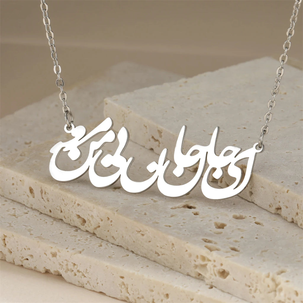A silver nameplate necklace with Arabic calligraphy script, displayed on a textured beige stone block background.