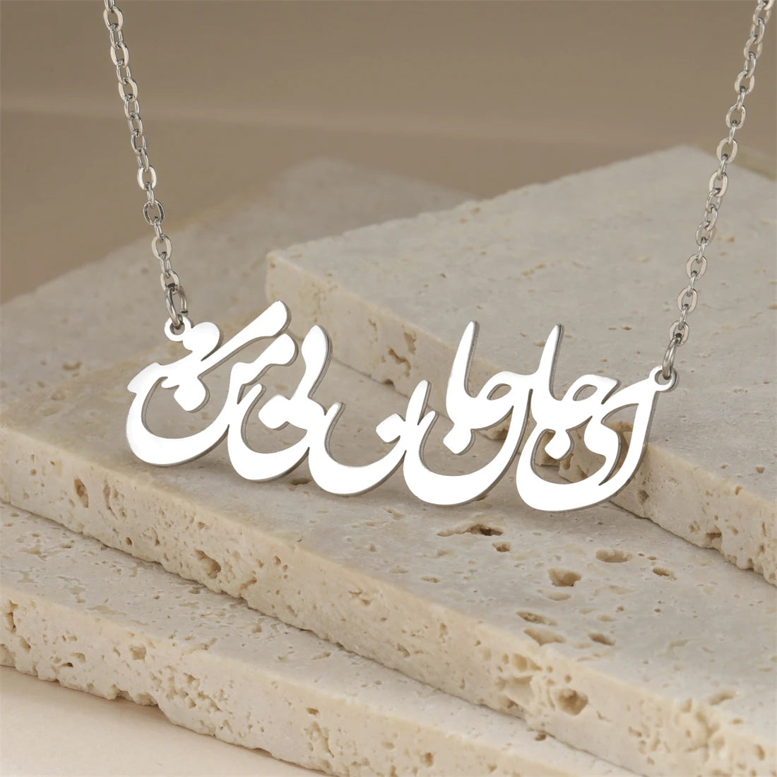 A silver nameplate necklace with Arabic calligraphy script, displayed on a textured beige stone block background.