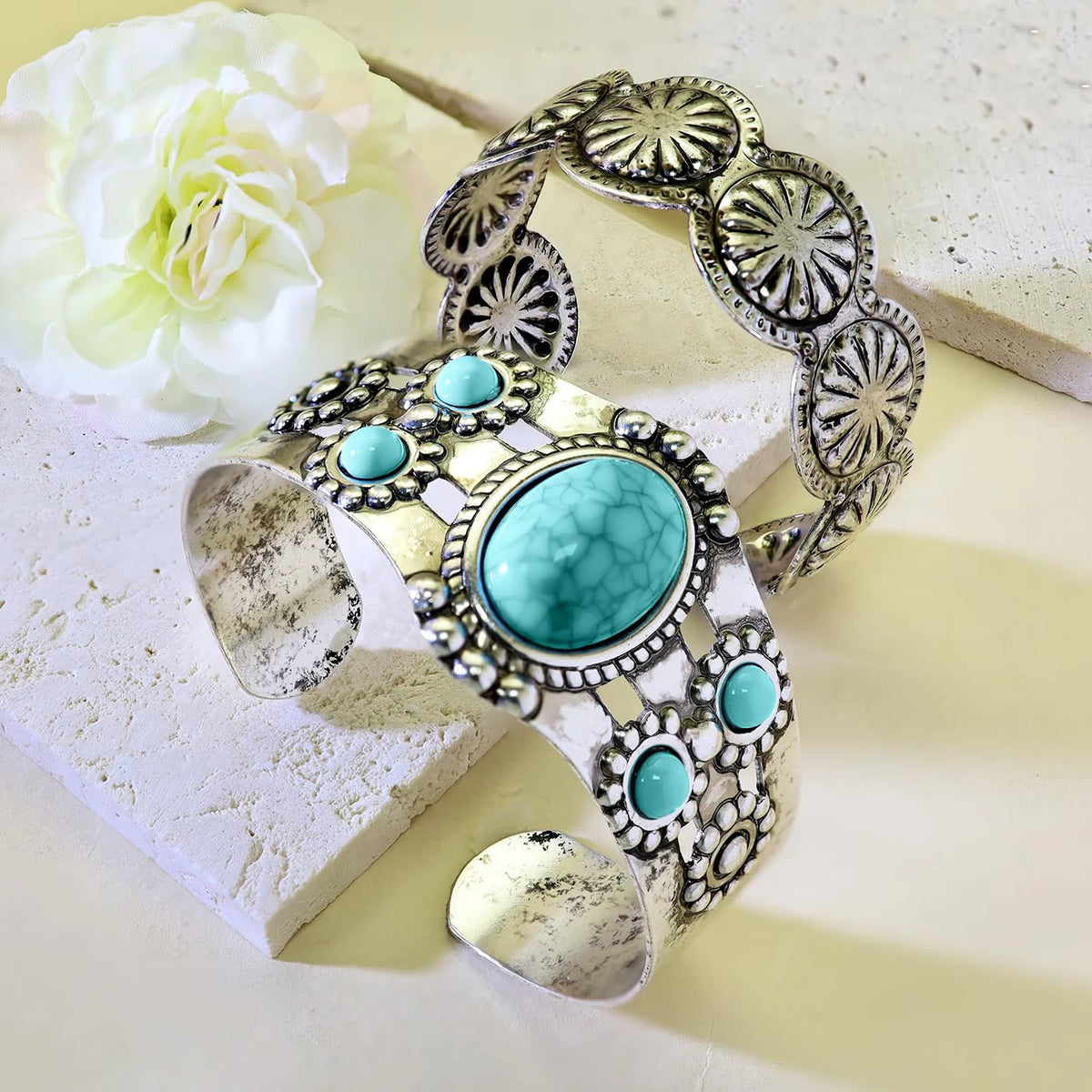 An ornate silver-toned cuff bracelet with a large oval turquoise center stone and several smaller round turquoise stones, styled on a light surface with a white flower and textured stone blocks.