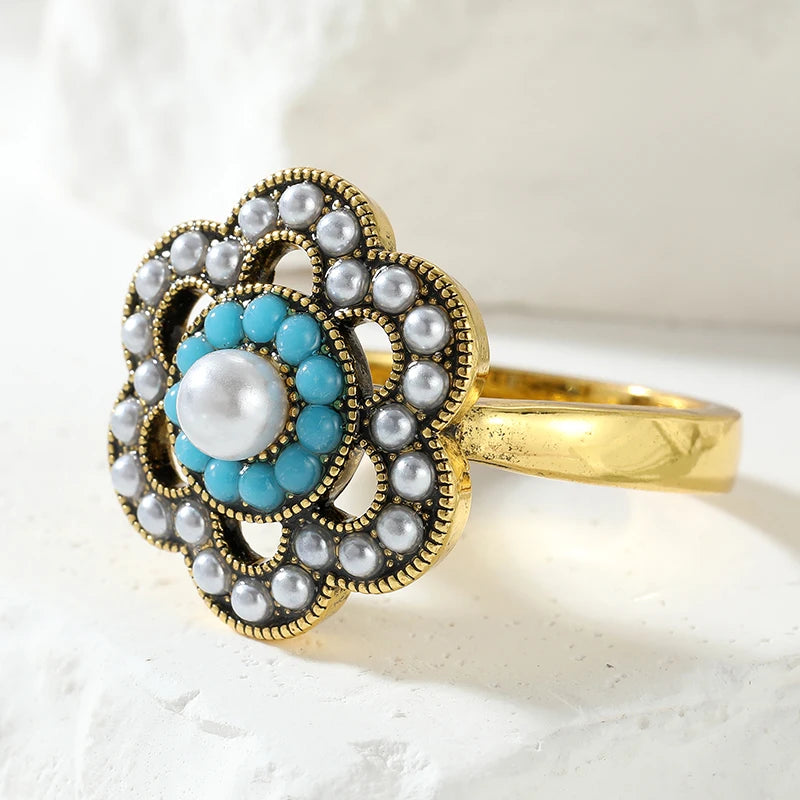 An ornate vintage-style ring with a gold-tone band and a floral cluster setting. The center features a large white pearl surrounded by a ring of bright turquoise blue stones, with multiple smaller white pearls and silver-toned beaded accents around the outer petals, set against a textured white background.