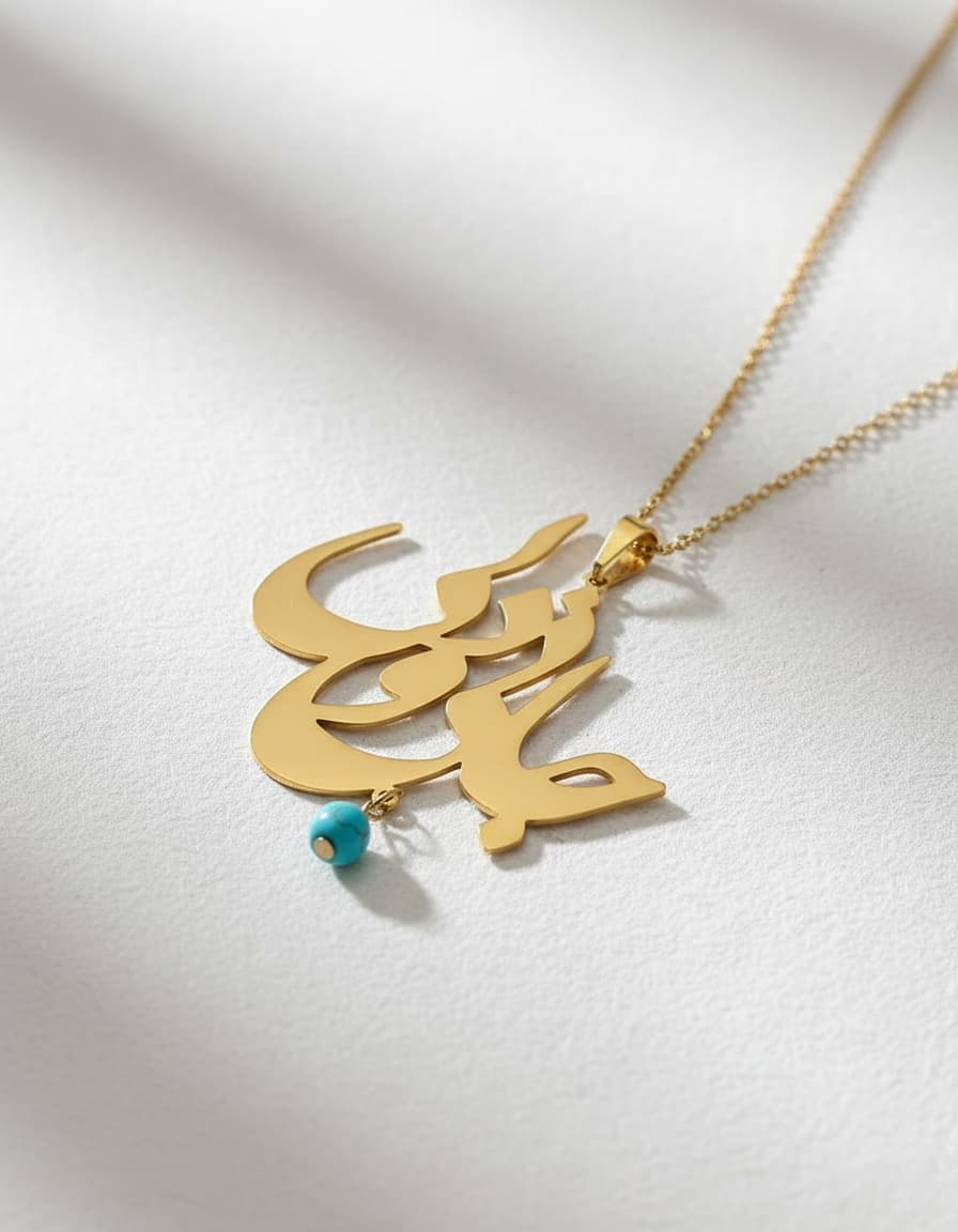 Gold-tone Persian calligraphy necklace with a dangling turquoise-blue bead on a white textured surface.